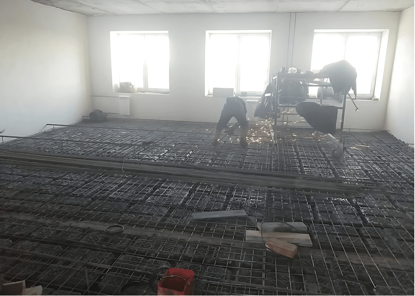 Workers performing welding over a reinforced concrete slab with visible void formers installed for floor re-leveling during building renovation.