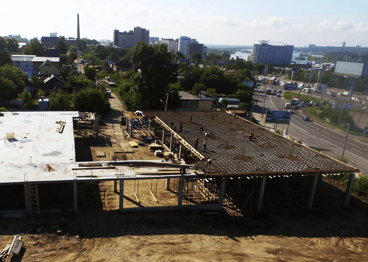 Aerial view of a construction site with workers installing reinforcement over a large slab framework, surrounded by urban roads and residential buildings in the background.