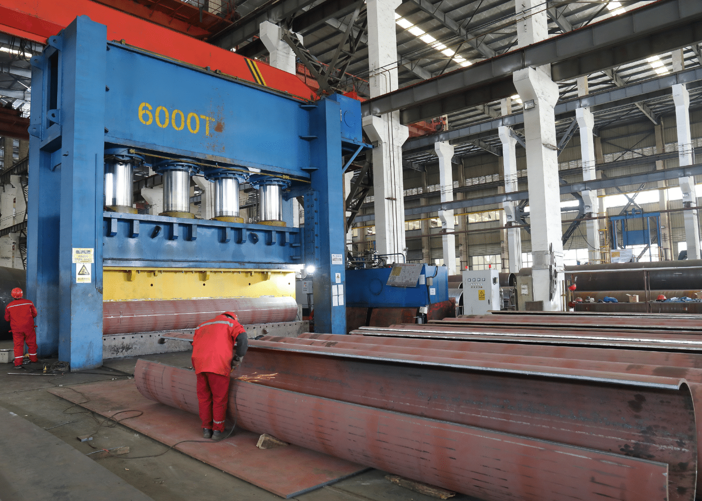 Industrial steel pipe forming line with workers and mechanical engineers operating a heavy-duty hydraulic press, forming a pipe from a steel plate.