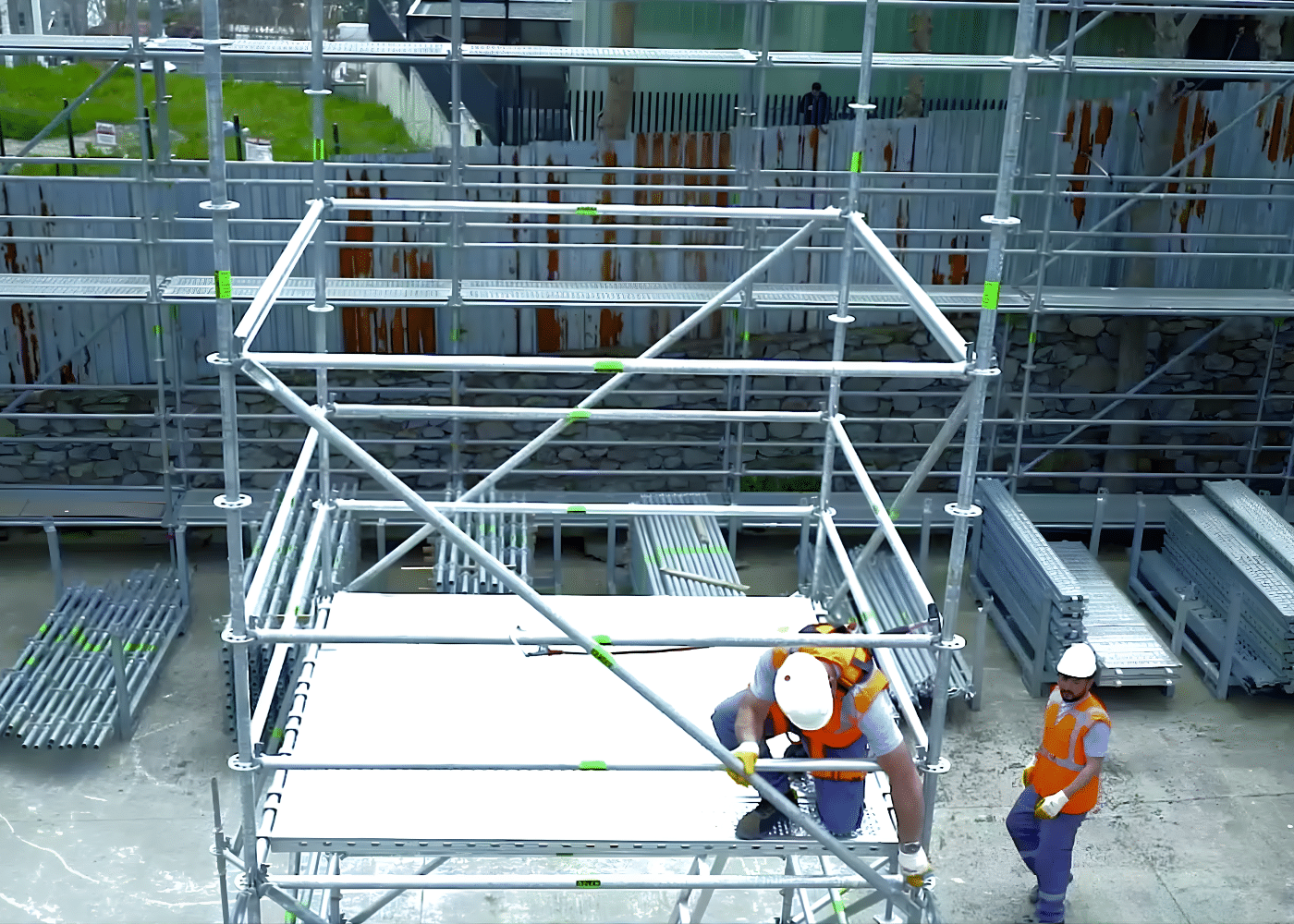 Construction workers are assembling a modular scaffolding tower on a job site, installing cross-braces and platforms by hand to ensure a stable, reliable work system.